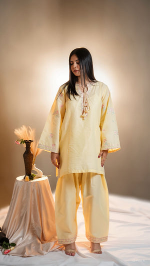 Woman in a light yellow traditional outfit standing in a softly lit room with a vase and flowers on a table.