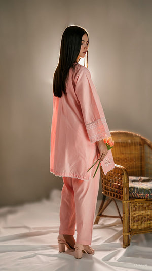 Woman in a pink outfit holding flowers, standing in a softly lit room with a wicker chair.