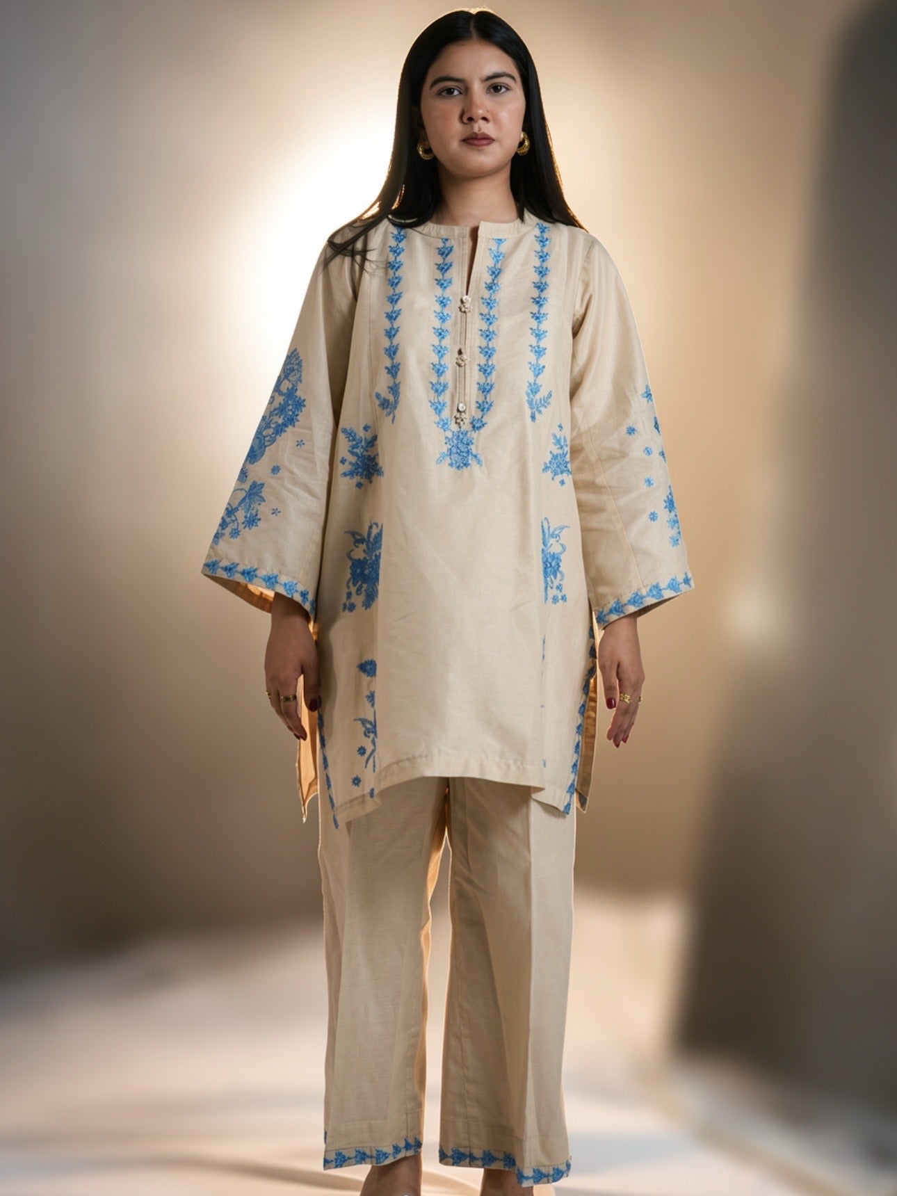 Woman wearing a beige outfit with blue embroidery against a neutral background