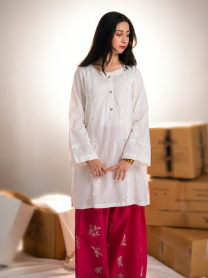 Woman wearing a white top and red pants standing among cardboard boxes.
