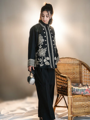 Woman in black embroidered jacket and pants standing next to a wicker chair with a patterned cushion.